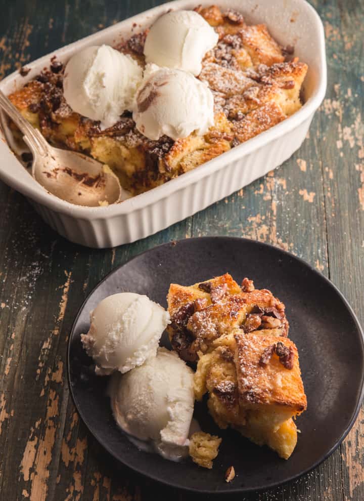 bread pudding in a white baking dish and a piece on a black plate with 2 scoops of vanilla ice cream