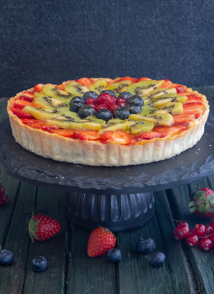 Fruit flan on a black cake stand, with fresh fruit around it.